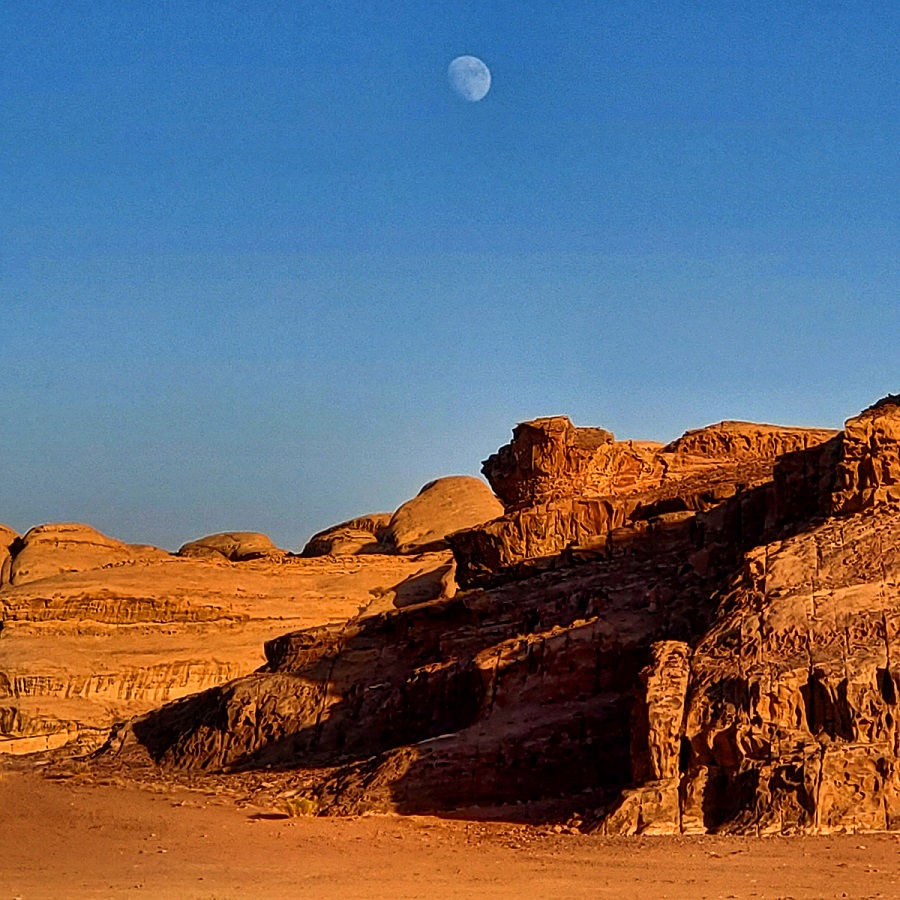 Red sand dunes, sandstone rises from it. A full moon rises above it in the still blue sky.