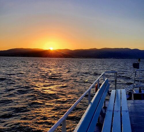 The sun sets over the island of Chios from the deck of the incoming ferry. The water is a deep blue with only mild small waves.