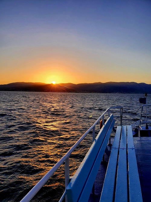 The sun sets over the island of Chios from the deck of the incoming ferry. The water is a deep blue with only mild small waves.