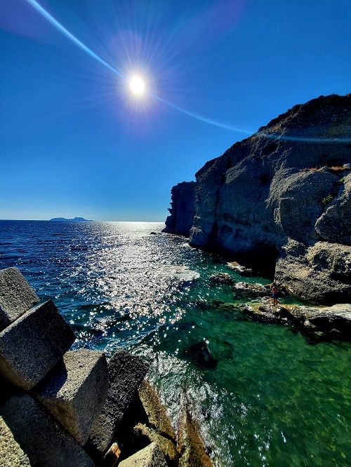 The rocky steep cliffs at on the shore of Crete. The water is aqua and deeper blue further from shore. The sun is bright and creates an arc in the camera's lens. The concrete break wall is just visible in the lower left portion of the photo.