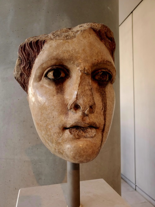 The sculpted face of a woman in stone. Grey streaks run beneath both eyes down her cheeks. Ages in the weather have given her the appearance of her make up running.