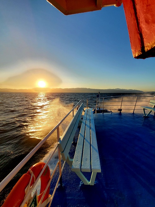 Waves splash up from the blue painted hull of the ferry from Turkey to Chios, Greece. The sun is beginning to set and is low on the horizon, shining through the spray.