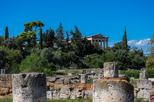 Roman ruins stand on a hilltop surrounded by cypress and cedar trees. Columns stand in the foreground among the walls of ruins.