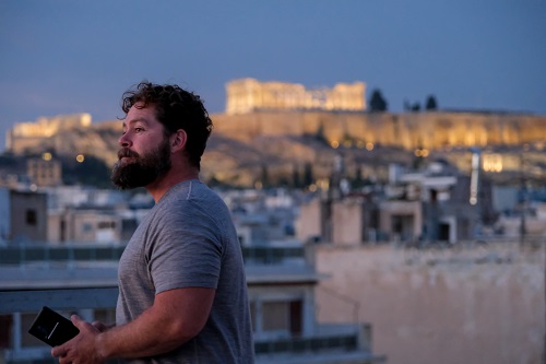 Michael Stahl standing on the terraza of a rental apartment in Athens. The Acropolis covers the hill in the background, buildings in Athens can be seen below it.