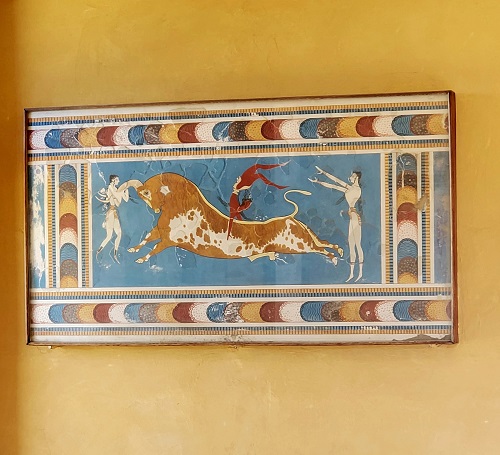 A yellow plastered wall in Knossos. A painting of a charging bull with a man throwing another on its back and another fighting it by the horns.