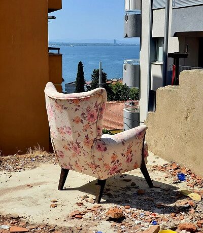 A wingback chair upholstered in white with roses looks out from the ruins of a house. The walls have fallen away giving a perfect view of the ocean from the hilltop above. Cigarette butts litter the ground with a yellow bowl and bottlecaps among other trash.