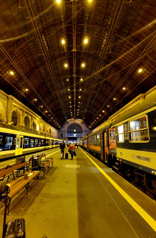 The arched roof of Bucharest station far overhead of travellers walking toward the station. Amber light makes everything lends everything a yellow tone. A small white circle of light is high on the wall of the station overhead. The station clock.