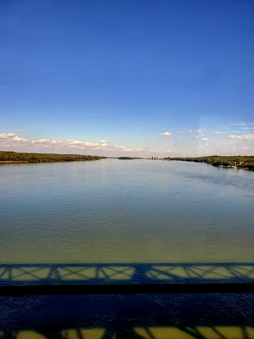 The shadow of the train trestle is cast onto the river below. The banks are lined with trees, many still green but with yellowing leaves. the river is a shade of mossy green.