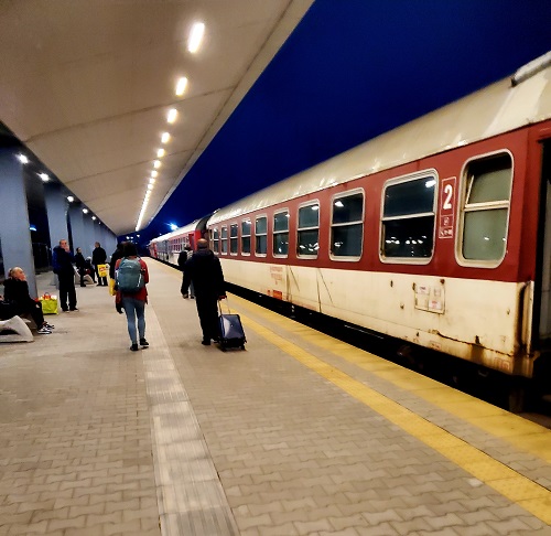 A white and red train sits with dark windows next to a platform where a few people sit. Rust spots and streaks of dirt cover the train.