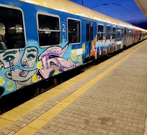 A train with dark windows sits at a platform in Sofia, Bulgaria. the sides are tagged with graffiti, spots of rust and streaks of dirt mar the paint.