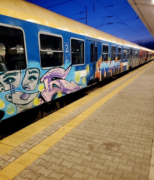 A train with dark windows sits at a platform in Sofia, Bulgaria. the sides are tagged with graffiti, spots of rust and streaks of dirt mar the paint.