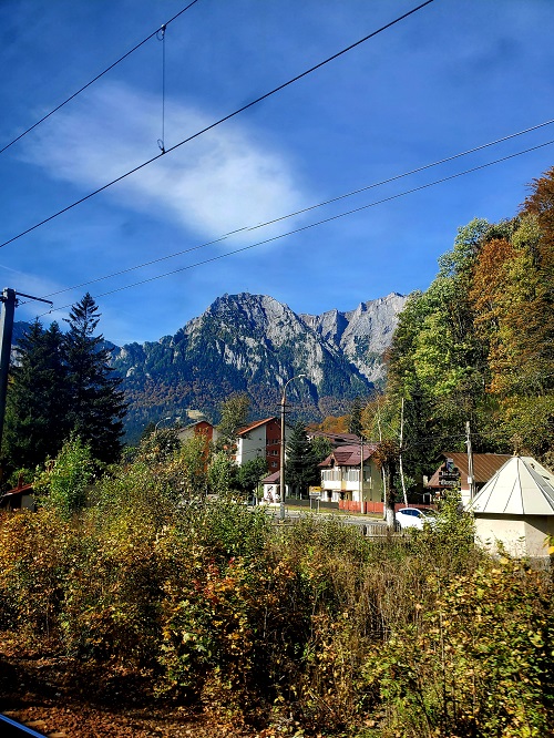 The grey stone mountaintops of the Carpathians seen over the tops of mountain chalet tiled roofs. Pine and other hardwood trees cluster around them, some leaves are changing to shades of orange. The sky is bright blue with light clouds above.