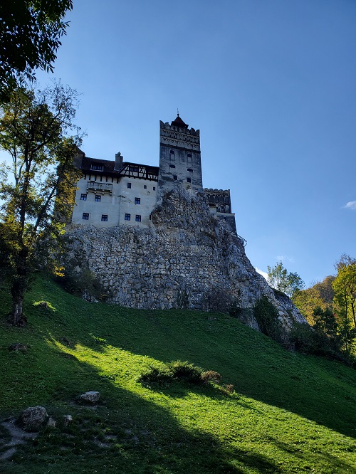 Castle Bran covers a hilltop surround by deep green grass with layers of stone block. White plastered towers rise above it. The stone tiled roof many stories from the grass below. Small windows adorn the tower and the walls.