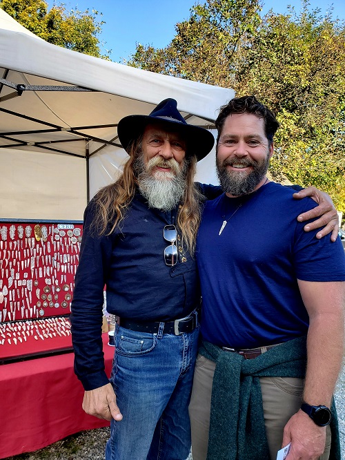 Michael Stahl and the artist and carver Manole stand together for a photo. Both with beards but, the carver's hair is shoulder length and he wears a fedora-style hat.