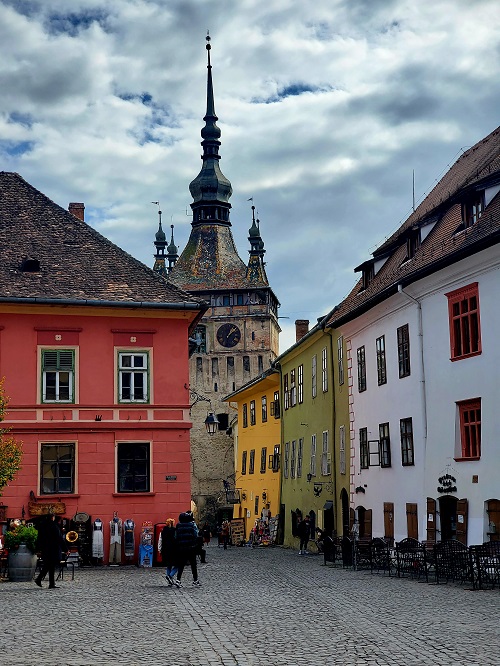 Baroque style buildings sit on either side of a narrow street. At the end a clock tower's spire rises higher than any other rooftop.