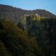 The walls and ruins of Peonari castle cover the top of a ridge seen from a distance. Treetops come to the base of its walls. Surrounded by greens and oranges of autumn.