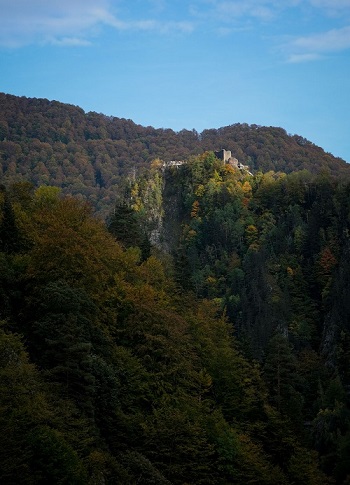 The walls and ruins of Peonari castle cover the top of a ridge seen from a distance. Treetops come to the base of its walls. Surrounded by greens and oranges of autumn.