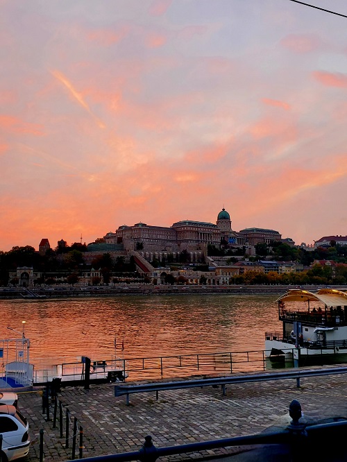 A bright sunset is reflected in the Danube. The Palace sweeps across the riverbank above.