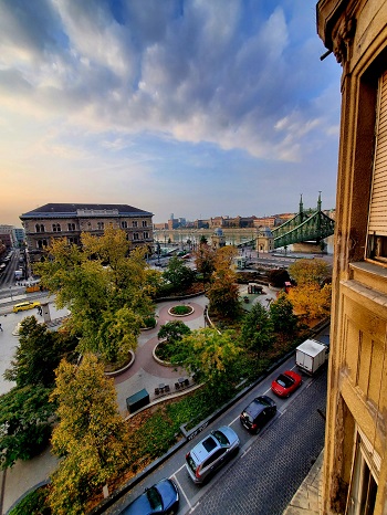 The sun beginning to rise over the buildings surrounding the open square. The Liberty Bridge, a seafoam green over the wide Danube with the clouds above.