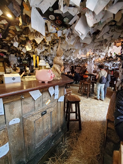 Paper notes hang from the wooden ceilings of the For Sale Pub. Straw covers the floor and candles light each table.